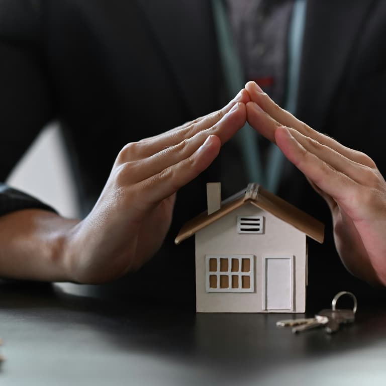 Hands forming a protective gesture over a small house model, symbolizing home security and insurance